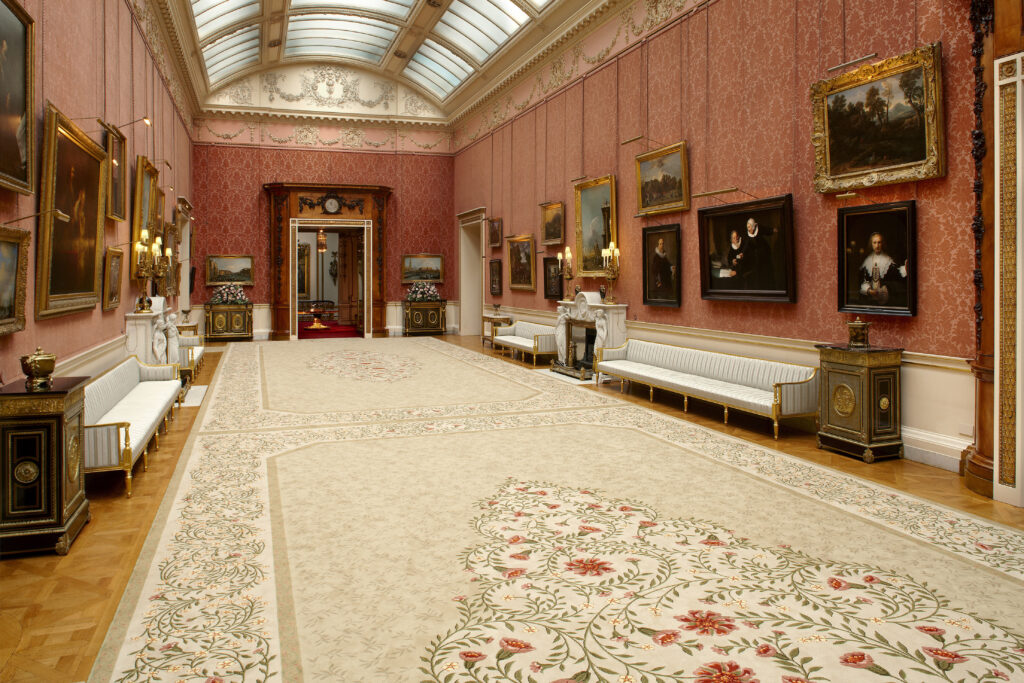 An image of a cream carpet with red floral design, set in the picture galelry of Buckingham Palace, a long, grand room with rose pink walls adorned with old master paintings.