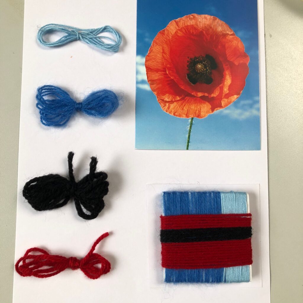 A4 paper with a picture of a red poppy with a black centre, accompanied by small samples of yarn. A square of foamboard is wrapped in yarn reflecting the colours of the poppy.