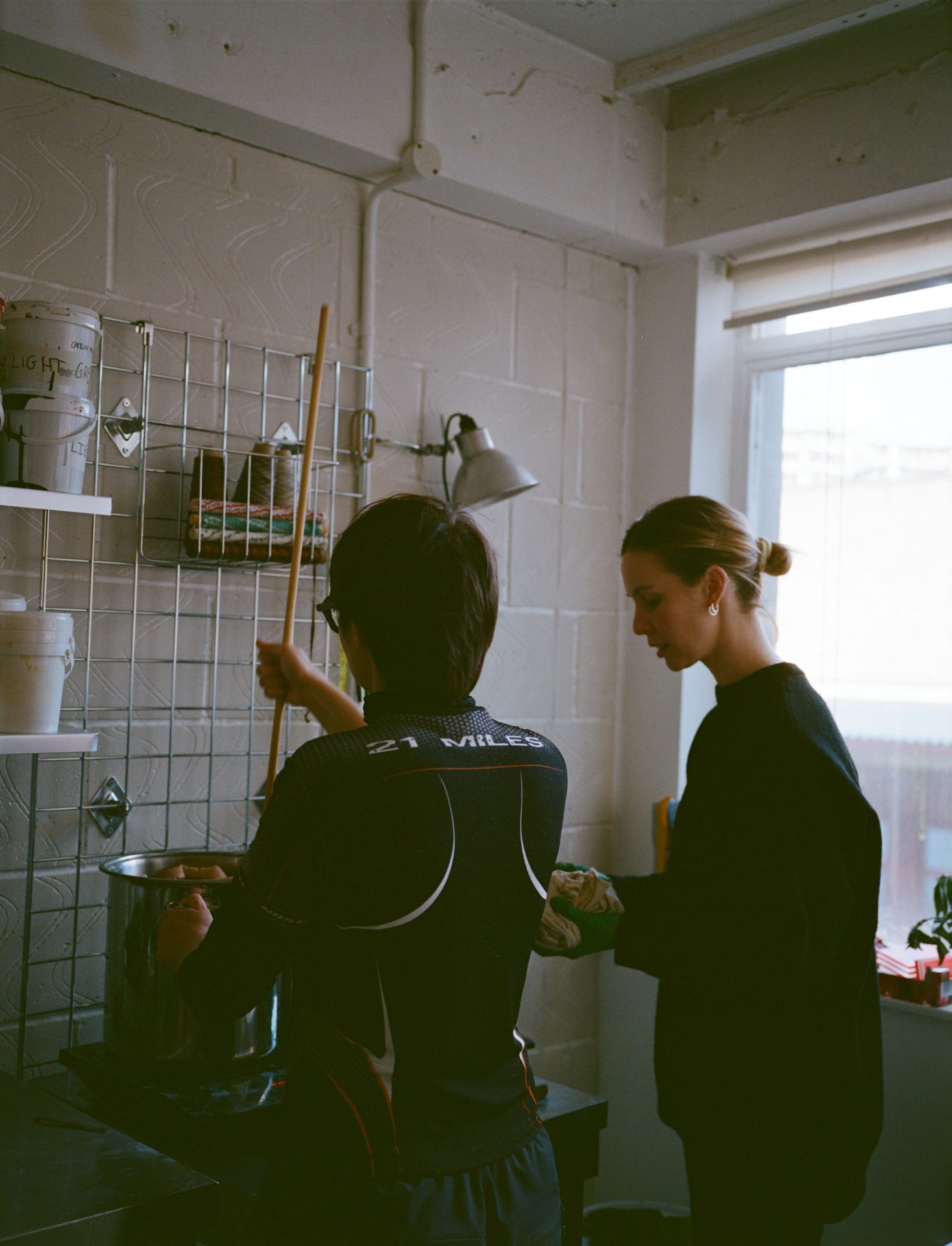 Two workshop participants with backs to camera stir dye pots and chat at a London natural‑dye session.