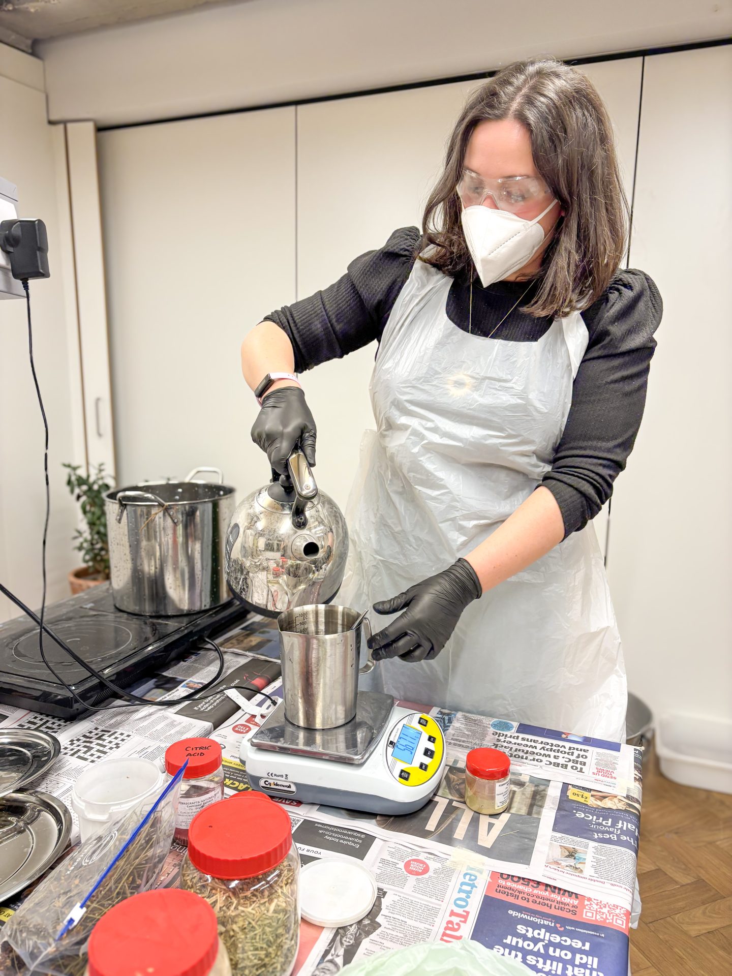 Workshop attendee wearing mask and glasses pours water into a dye pot during natural dye session in London.