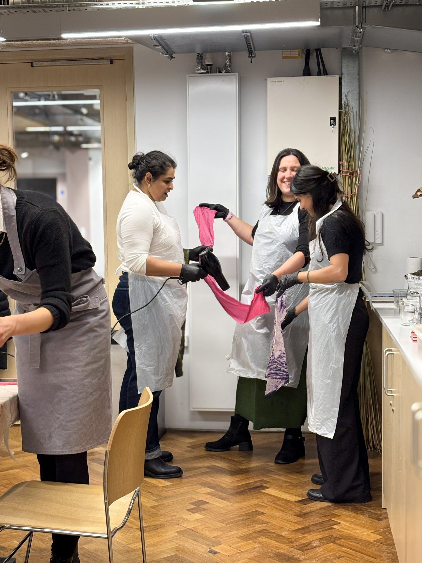 Three people at a natural‑dye workshop in London use a hair dryer to speed‑dry a freshly dyed scarf.