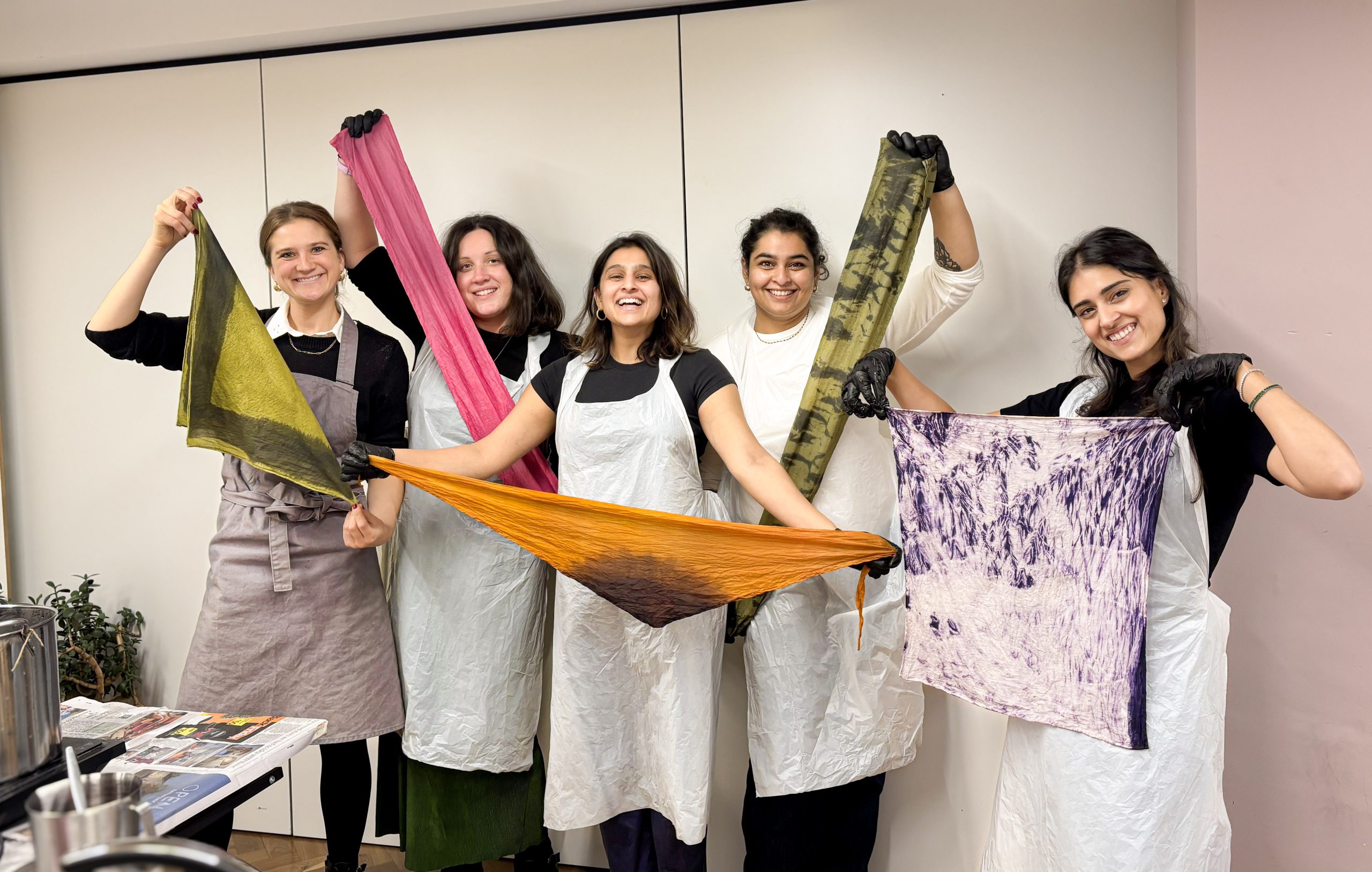 Five smiling participants at a natural dyeing workshop in London proudly show their naturally dyed bandanas of green, pink, orange, tiger‐print and purple shibori styles.