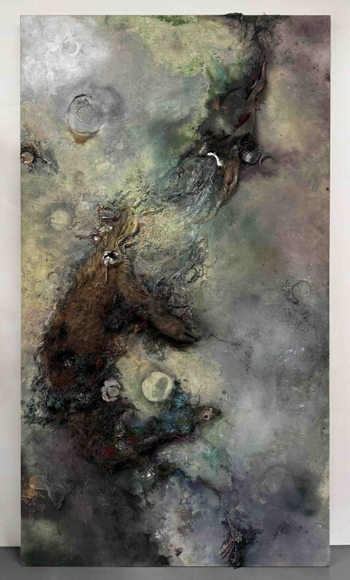 “Large sculptural wall painting with a dark, textured surface combining human hair, deer taxidermy, plastic waste and reflective materials.”